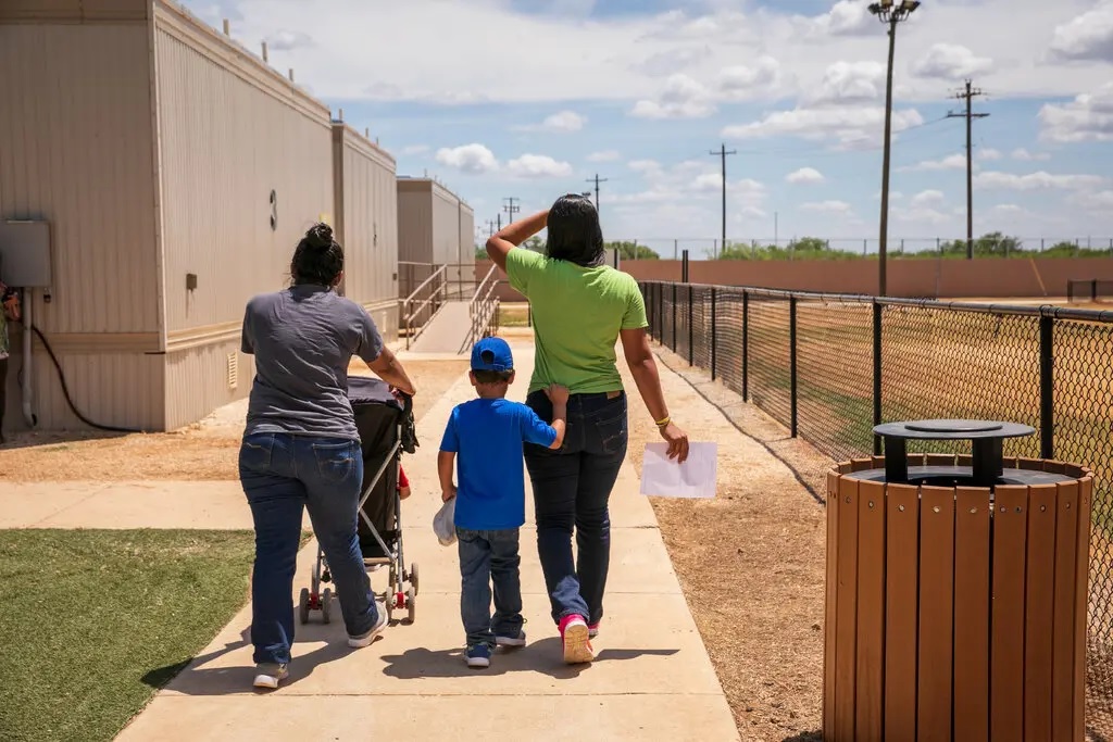 A woman pushing a stroller walking with a woman and a child, seen from behind, next to several trailers and a low fence.