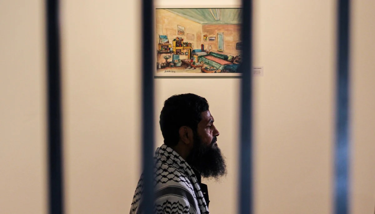 Ahmed stands before a painting he made portraying his room in Guantanamo Bay with rails of IVS art gallery in the foreground.  Photo by Hassaan Ahmed