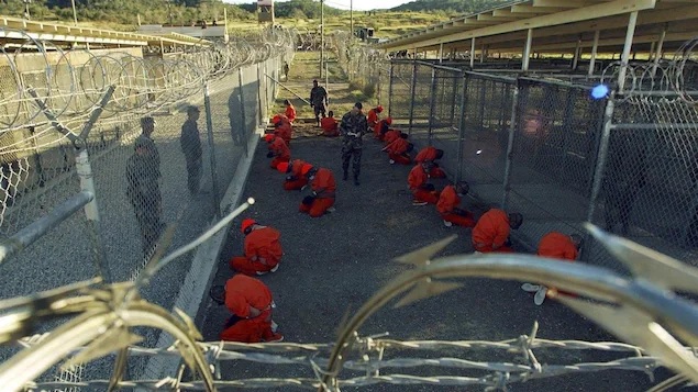 Detenidos vestidos con uniformes naranjas, estn esposados y agachados en el suelo de un recinto exterior cerrado por altas vallas con alambre de espino. Hay varios soldados que los vigilan.